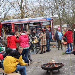 Ansammlung von Menschen um eine Feuerschale. Einige grillen etwas über der Feuerschale - andere stehen an einem Tisch und essen etwas, wieder andere stehen in der nähe und unterhalten sich.