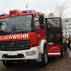 Ein großes Feuerwehrauto steht im Fokus. Rechts neben dem Auto steht ein Feuerwehr man der Zwei Kindern etwa sam Auto erklärt