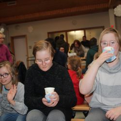 3 Kinder sitzen auf einer Bank, zwei davon trinken etwas, das andere Kind unterhält sich mit jemanden der nicht im Bild ist. im Hintergrund ist ein buntes Getümmel.