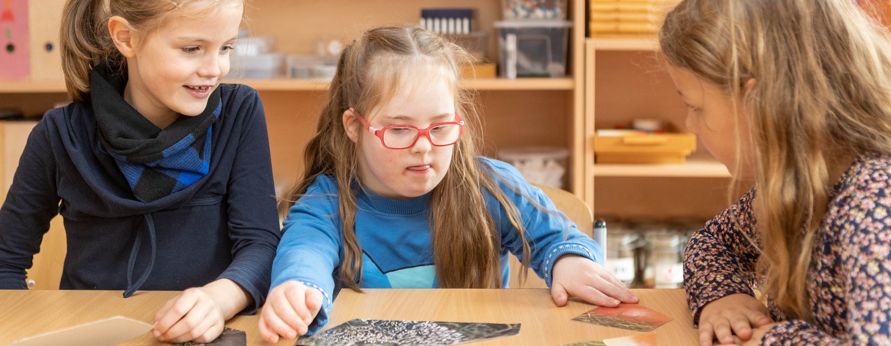 Zwei Kinder sitzen an einem Tisch während eine Lehrerin ihr etwas erzählt